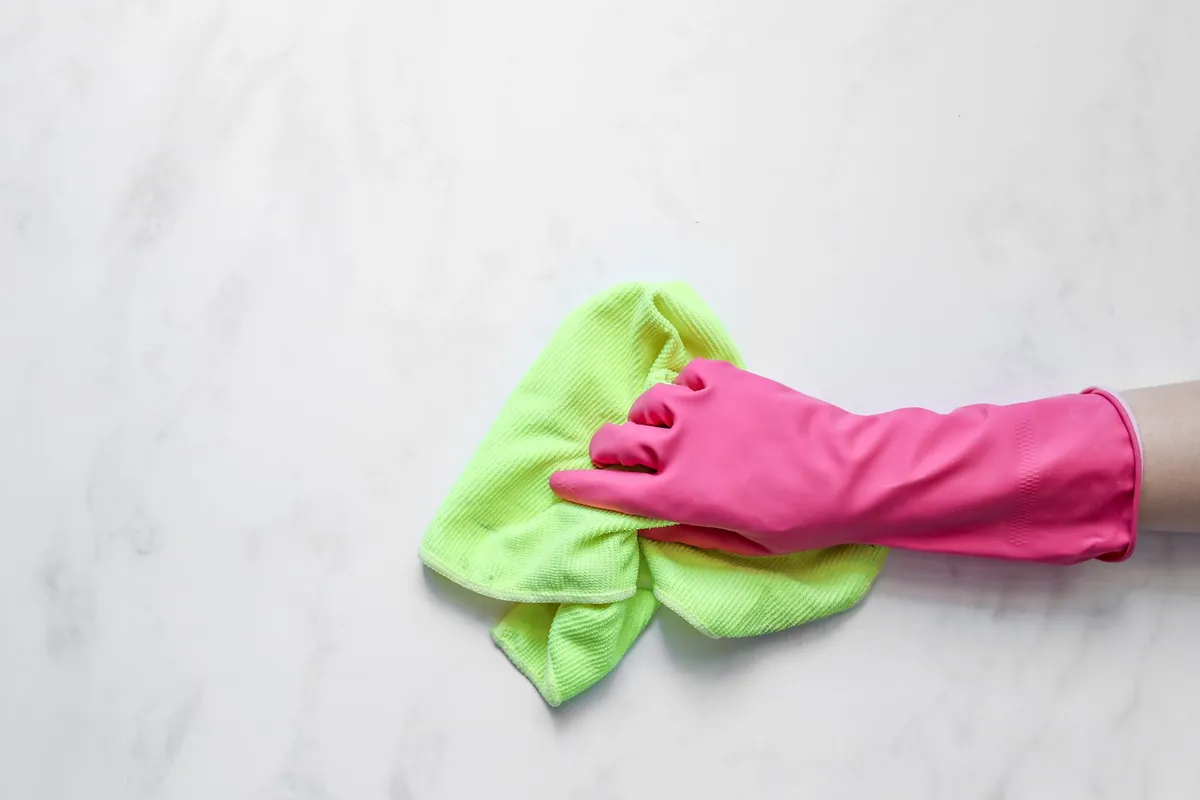 A hand in a pink rubber glove wiping a white marble counter with a green microfiber cloth
